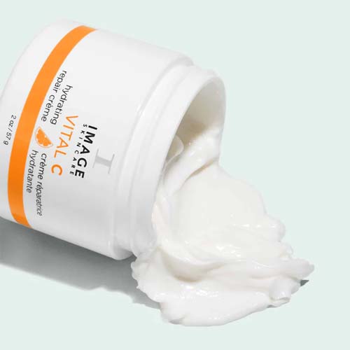 Een geopende pot VITAL C hydrating repair crème die op zijn kant ligt, waarbij de volle witte crème naar buiten komt op een lichte ondergrond.
