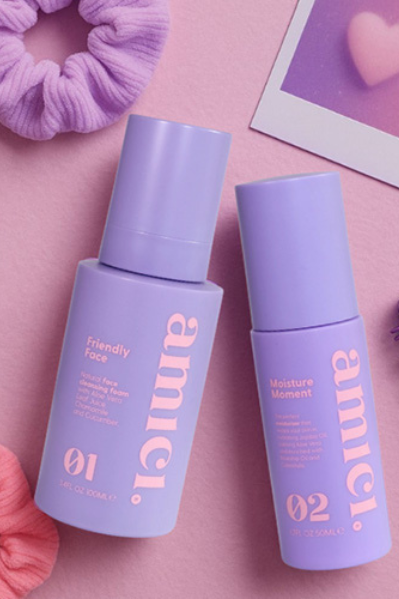 Twee paarse flacons van Amici Skincare, de Friendly Face cleansing foam en Moisture Moment moisturizer, liggend op een roze achtergrond met accessoires.