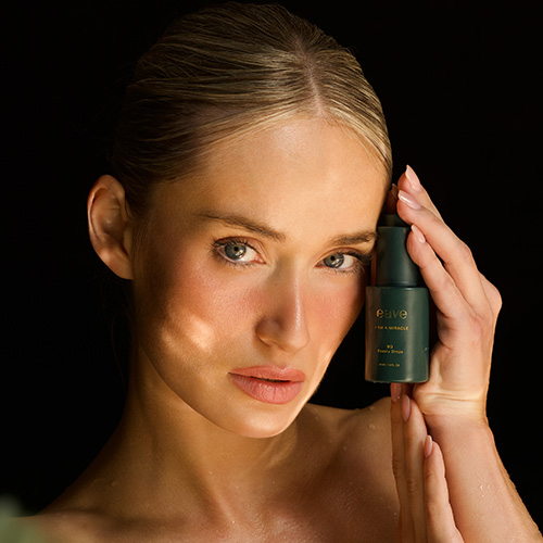 Een vrouw met een stralende huid houdt het donkergroene EAVE Beauty Drops flesje tegen haar gezicht. Warme belichting benadrukt de natuurlijke glans van haar huid.