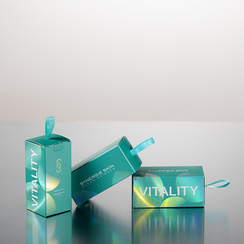 Drie groene Synergie Skin Vitality verpakkingen op een reflecterend oppervlak waarvan er één rechtop staat met het woord VITALITY erop en twee gekanteld liggen met zicht op het merkwachtwoord.