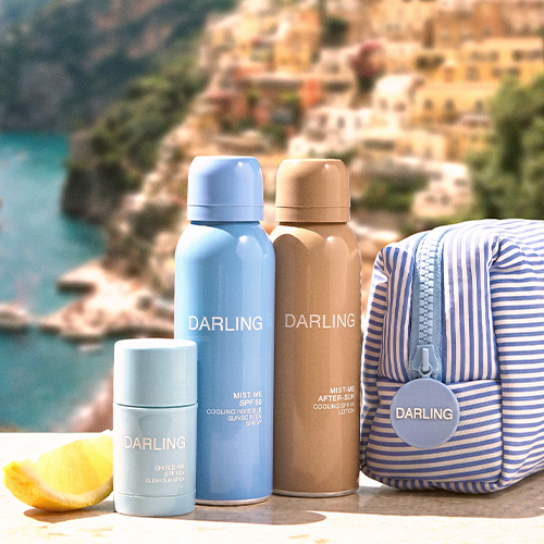 De zonnebrandproducten en het gestreepte tasje uit de Darling La Vacanza set sfeervol opgesteld in de zon met een Italiaans kustdorpje op de achtergrond.