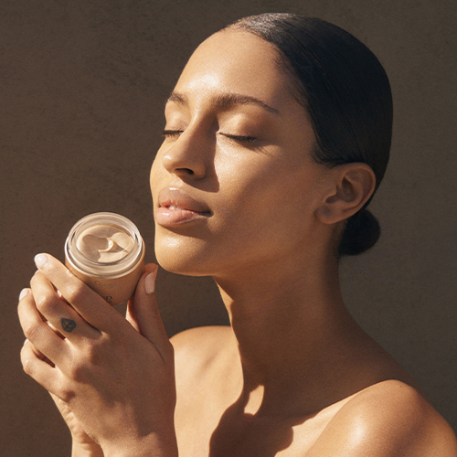 Een vrouw houdt een geopend potje Natura Bissé Diamond Cocoon Sheer Cream vast bij haar gezicht in warm zonlicht.