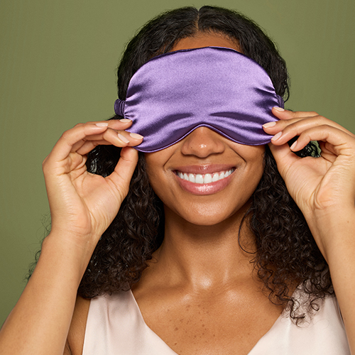 Lachende vrouw met donker krullend haar die een paars zijden slaapmasker voor haar ogen vasthoudt