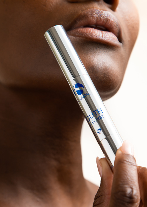 Een close-up van een hand die een zilverkleurige tube iS Clinical Youth Lip Elixir vasthoudt bij de mond van een persoon.
