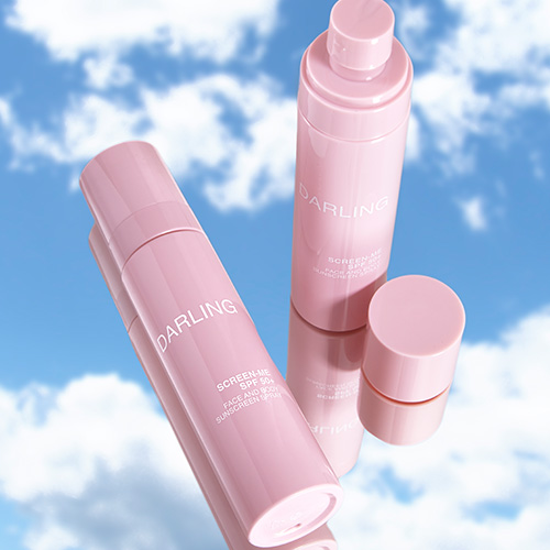 Roze DARLING SCREEN-ME SPF 50+ sprayflacons gefotografeerd op een spiegelende ondergrond met een reflectie van de blauwe lucht en wolken.