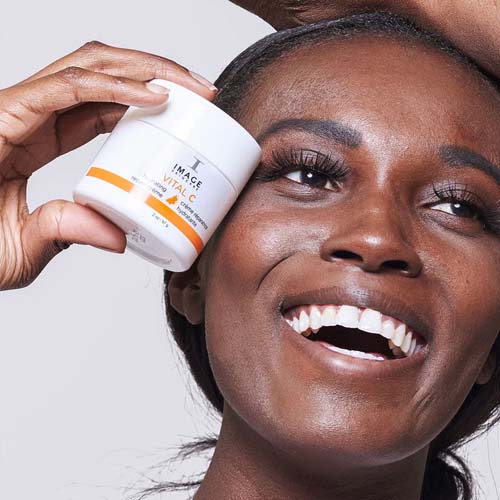 Een glimlachende vrouw houdt een pot IMAGE Skincare VITAL C hydrating repair crème tegen haar wang.