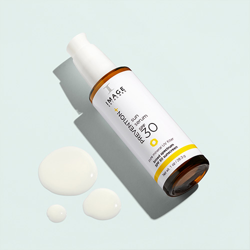 De flacon van het IMAGE Skincare Sun Serum SPF 30 liggend op een lichtblauwe achtergrond met drie druppels van het serum ernaast.