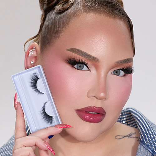 NikkieTutorials houdt de verpakking van de Nimya by NikkieTutorials wimpers vast. Ze draagt volledige make up, inclusief de wimpers in het doosje, roze blos en glanzende roze lippen. Ze heeft een denim jasje aan en een tattoo op haar sleutelbeen.