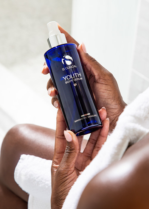 Persoon met donkere huidskleur houdt een blauwe fles iS Clinical Youth Body Serum vast, met een witte handdoek op de achtergrond.