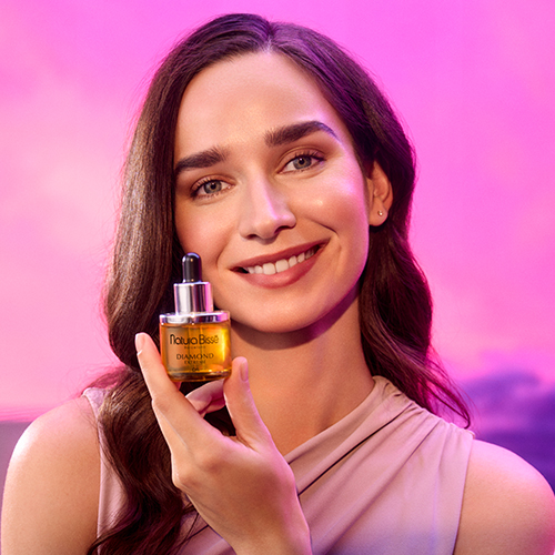 Een glimlachende vrouw houdt het flesje Natura Bissé Diamond Extreme Oil vast bij haar gezicht tegen een roze en paarse zonsondergang achtergrond.