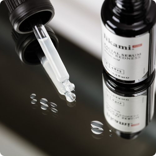 Een glazen flesje van het Likami Skin Redness serum met de pipet en enkele druppels vloeistof ernaast, weerspiegeld op een glanzende, donkere ondergrond. De afbeelding heeft afgeronde hoeken.