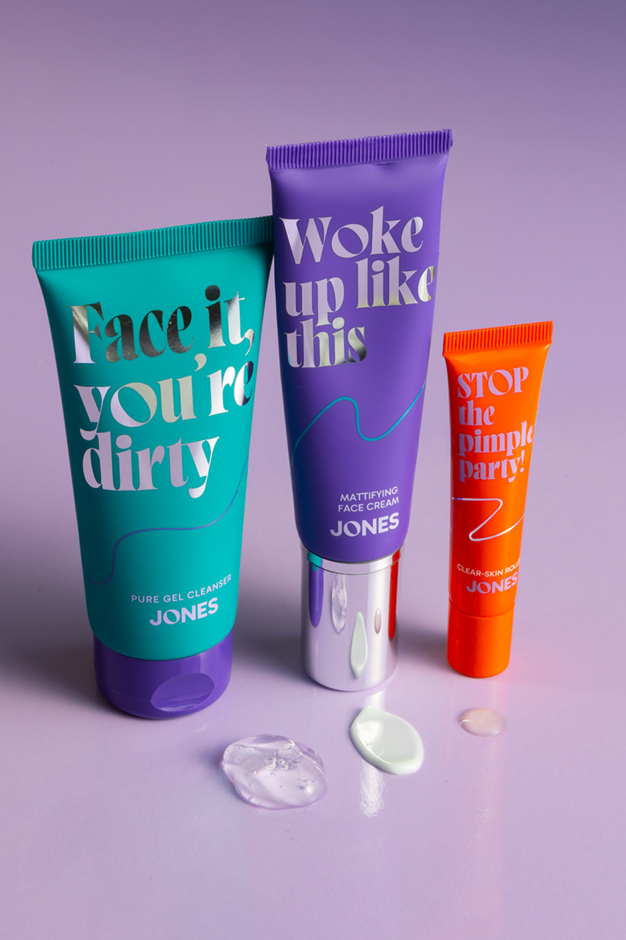 Drie kleurrijke tubes van Take It From Jones: de Face It You're Dirty gel cleanser, Woke Up Like This face cream en Stop The Pimple Party roller, staand op een paarse achtergrond met druppels van de texturen.