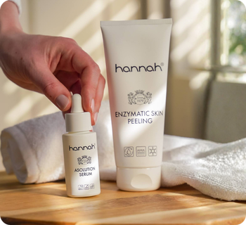 Een hand die het hannah asolution serum pipetflesje pakt dat naast een tube hannah enzymatic skin peeling op een houten tafel staat met een witte handdoek