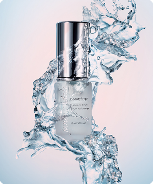 Een flacon Jane Iredale BeautyPrep Hyaluronic Serum omringd door dynamische waterspatten tegen een zachte, lichtblauwe achtergrond.