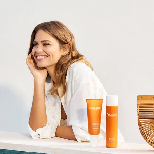 Een glimlachende vrouw in een witte blouse leunt op een witte muur met de Natura Bissé bodycream en een andere tube op de voorgrond.