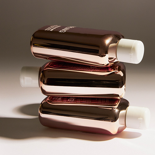 Drie koperkleurige flacons van Darling Soothe Me After-Sun Lotion horizontaal op elkaar gestapeld in een warme, sfeervolle setting met zachte schaduwen.