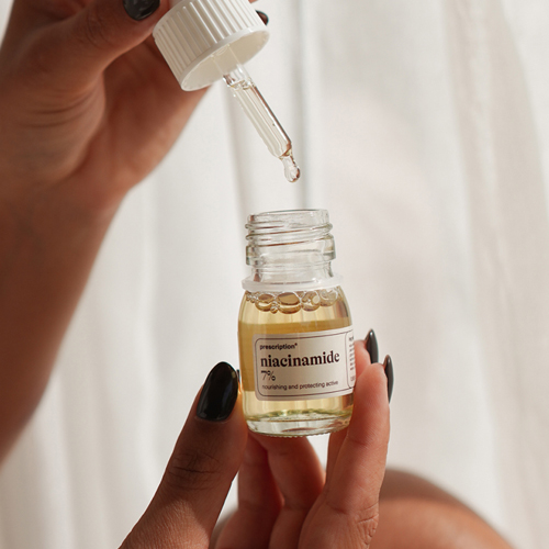 Een close up van een hand met zwarte nagels die een druppelaar boven een open glazen flesje Prescription niacinamide 7% houdt. De achtergrond is wazig en wit.