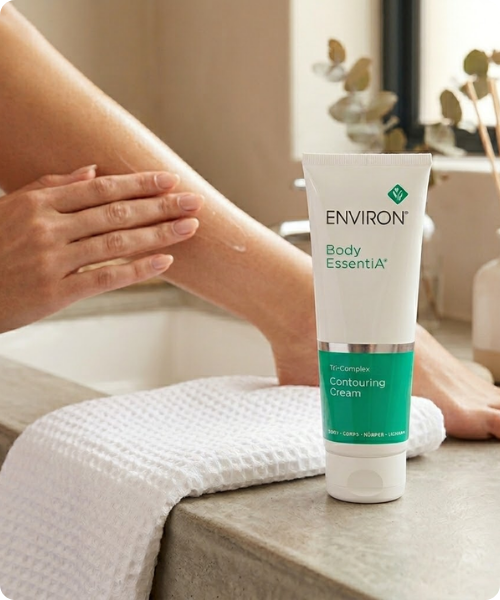 Een sfeerbeeld met afgeronde hoeken waarin een staande tube van de Environ Body EssentiA Tri-Complex Contouring Cream op een witte handdoek is geplaatst, met op de achtergrond een vrouw die haar been insmeert