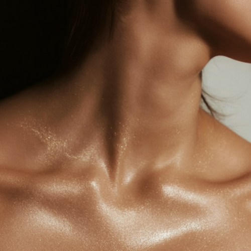 Close-up van de hals en het decolleté van een vrouw waarop de glinsterende gouden deeltjes van The Glow Elixir een subtiele, stralende glans achterlaten.