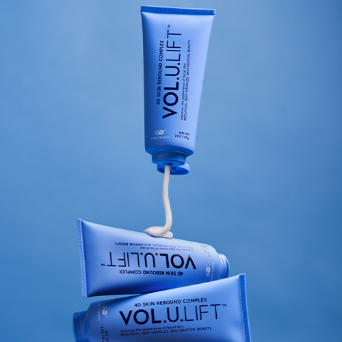 Drie blauwe tubes van IMAGE Skincare VOL.U.LIFT 4D Skin Rebound Complex waarvan er één witte creme op een andere tube spuit, weergegeven in een creatieve opstelling.