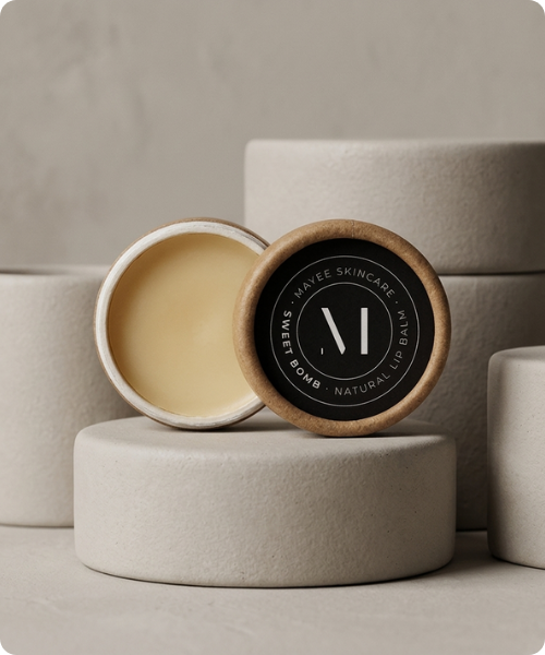 Twee ronde kartonnen potjes Mayee Sweet Bomb natuurlijke lippenbalsem op ronde beige sokkels een potje is open en toont de balsem het andere potje toont het deksel met het logo