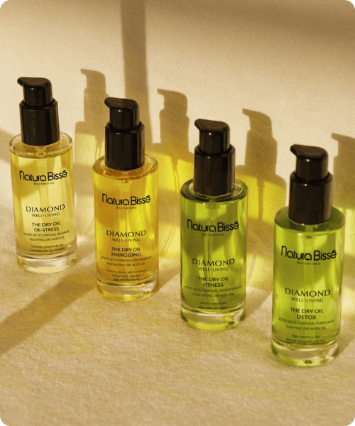 Vier verschillende flacons Natura Bisse Diamond Well Living droge lichaamsoliën De Stress, Energizing, Fitness en Detox opgesteld in een rij met zachte schaduwen.