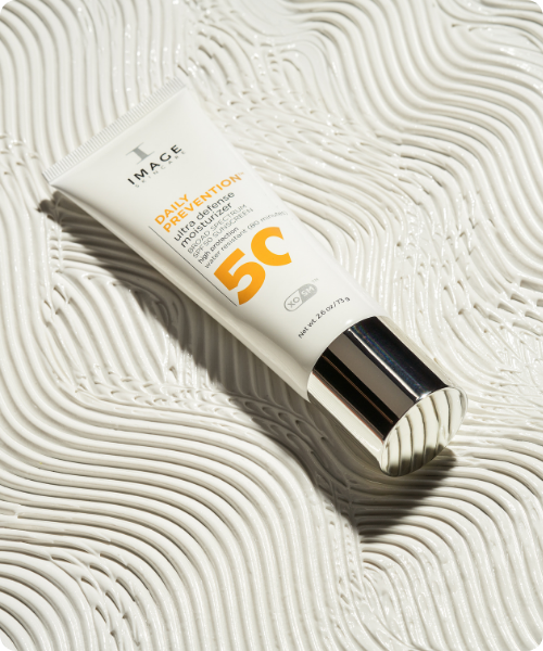 Een witte tube IMAGE Skincare Daily Prevention Ultra Defense Moisturizer met SPF 50, liggend op een artistieke ondergrond van dikke, golvende witte crème-texturen. De tube heeft een glanzende zilveren dop en gele details in de productnaam.