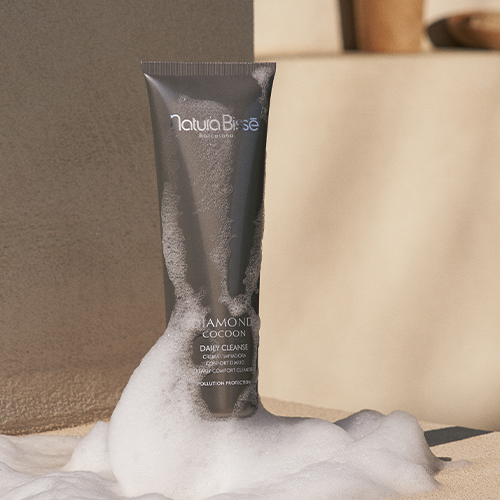 De Diamond Cocoon Daily Cleanse tube van Natura Bissé omringd door wit reinigingsschuim op een zandkleurige ondergrond.