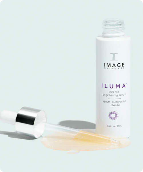 Een geopende witte flacon van het IMAGE Skincare ILUMA brightening serum met de pipet ernaast liggend in een plasje van het transparante, goudkleurige serum.
