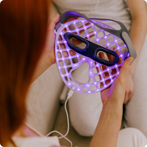 Een close-up van iemand die het rastervormige Likami LED-masker vasthoudt, dat oplicht met paarsblauwe lampjes. De afbeelding heeft afgeronde hoeken.