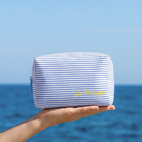 Een hand die het blauw met wit gestreepte Darling La Vacanza toilettasje omhoog houdt tegen een achtergrond van een helderblauwe zee en lucht.