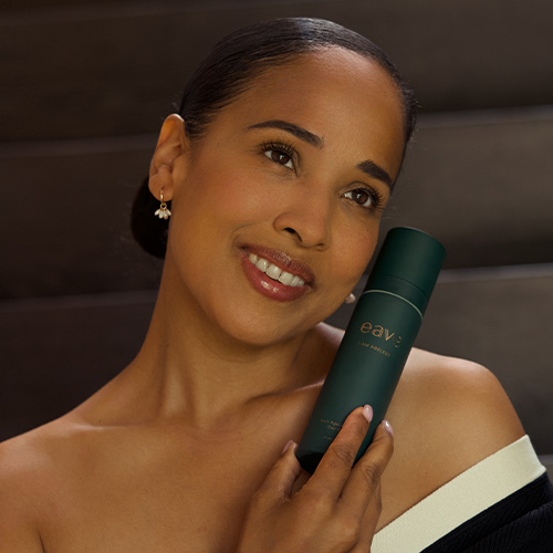 Een glimlachende vrouw die de donkergroene flacon van de EAVE I AM AGELESS dagcrème bij haar gezicht houdt.