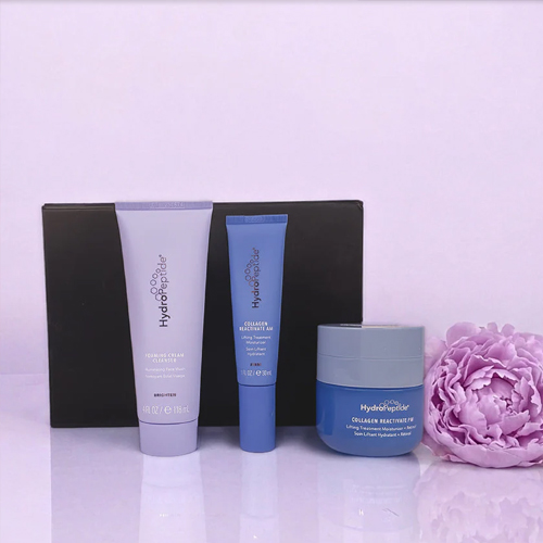Drie blauwe HydroPeptide producten uit de Glow With Love set gepresenteerd voor een zwarte geschenkdoos met een paarse bloem op de achtergrond.