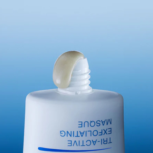Een close-up van een witte tube "Tri-Active Exfoliating Masque" waar een klodder van de grijzige, gelachtige exfoliant uit de opening komt tegen een blauwe achtergrond.