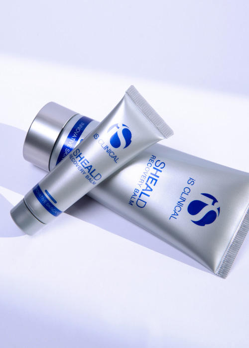 Twee zilverkleurige tubes iS Clinical Sheald Recovery Balm die speels op elkaar liggen tegen een lichte achtergrond.