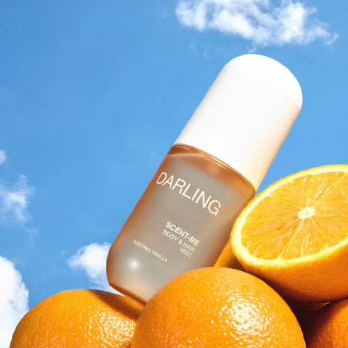 De capsulevormige flacon van de Darling Scent Me mist poserend tussen een stapel frisse sinaasappels tegen een strakke blauwe lucht met witte wolken