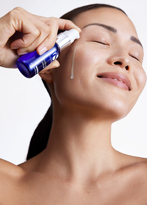 Een persoon met gesloten ogen die een druppel iS Clinical Hydra-Cool Serum op haar wang aanbrengt vanuit een blauw glazen flesje.