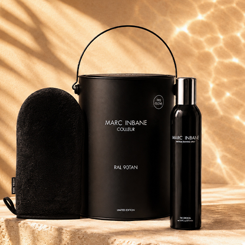 Sfeerbeeld van de Marc Inbane RAL 90TAN geschenkset met de Natural Tanning Spray fles een zwarte emmer en een tanning glove op een stenen ondergrond met waterreflecties op de muur