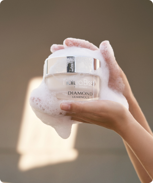 Twee handen die een pot Natura Bissé Diamond Luminous Cleanser vasthouden, bedekt met een dikke, zachte laag reinigingsschuim.