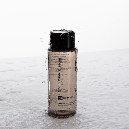 De fles Synergie Skin Micellution micellair water staat in een plas water terwijl er dynamische waterdruppels en spatten over de fles en dop heen vallen tegen een witte achtergrond.