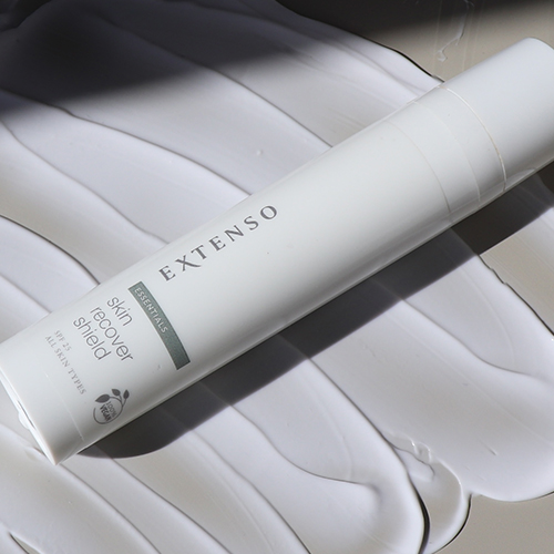 Een fles Extenso Skin Recover Shield die diagonaal op een ondergrond van uitgesmeerde witte crème ligt.