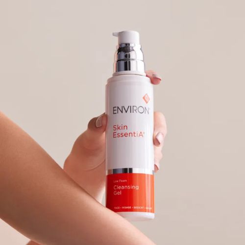 Een hand houdt de Environ Skin EssentiA Low Foam Cleansing Gel flacon vast tegen een egale, beige achtergrond.