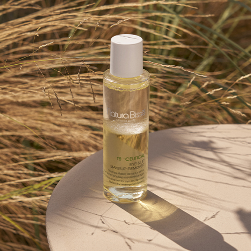 De Natura Bissé Eye & Lip Makeup Remover op een ronde stenen tafel buiten, omringd door wuivend riet en verlicht door warm, natuurlijk zonlicht.