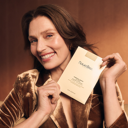 Een lachende vrouw met bruin haar, gekleed in een fluwelen jasje, houdt een goudkleurige verpakking van Natura Bissé Essential Shock Intense Instant Lift Mask vast met beide handen. Ze kijkt naar de camera. De achtergrond is een warme, koperachtige muur.