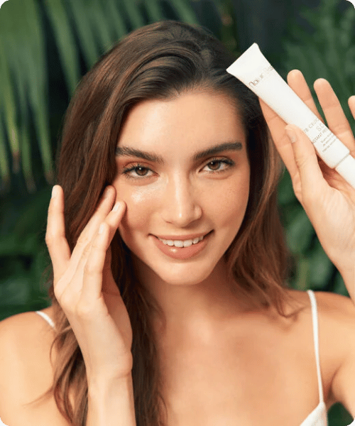 Een vrouw met een stralende huid houdt een tube Natura Bissé Tolerance SOS Instant Rescue vast bij haar gezicht, met een groene achtergrond van bladeren.