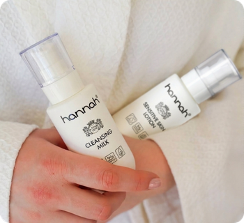 Een close up van een persoon in een witte badjas die twee hannah gezichtsverzorgingsproducten vasthoudt, de Cleansing Milk en de Sensitive Skin Lotion.