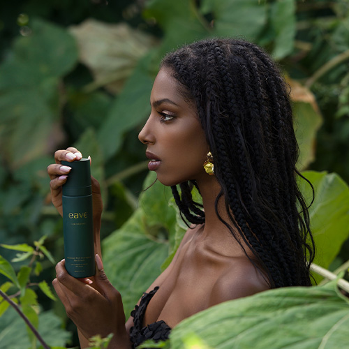 Een model houdt de Eave I AM RADIANT Day Cream vast tegen een achtergrond van grote, groene bladeren. Ze draagt een zwarte top met ruches en goudkleurige oorbellen. Haar haar zit in vlechtjes.