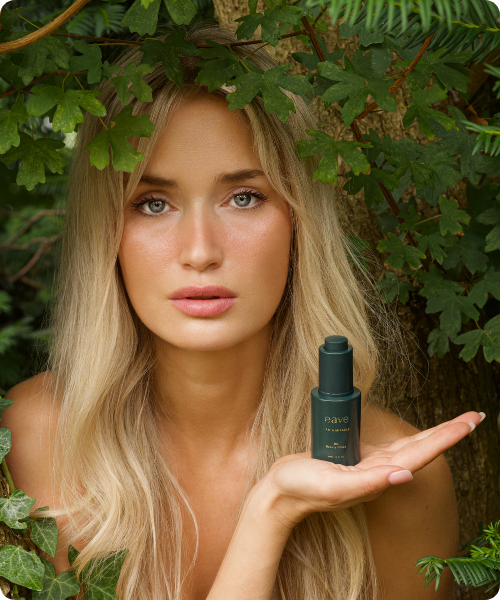 Een vrouw met lang blond haar staat in een natuurlijke omgeving met groene bladeren en presenteert een klein, donkergroen flacon van 'Eave I Am A Miracle' op de palm van haar hand.