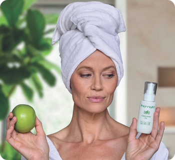 Een vrouw in een witte badjas houdt een groene appel en een flesje hannah cleansing oil omhoog