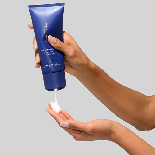 Hand van een persoon die een blauwe tube HydroPeptide Exfoliating Cleanser vasthoudt en reinigingsschuim op de handpalm doseert.
